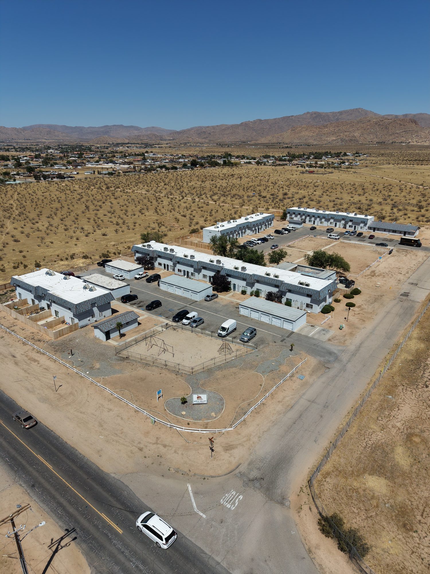 Aerial view looking north over Apple Valley Townhomes toward the mountains: the townhome buildings and garages in the foreground, Sioux Road along the left, and the town of Apple Valley spread across the desert floor behind.