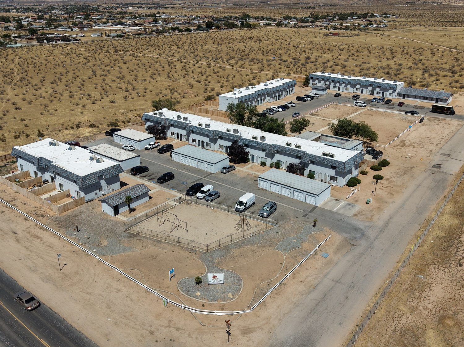 Aerial view of Apple Valley Townhomes: ten low two-story buildings with grey tile roofs around a paved lot, the garages and playground at the front along Sioux Road, fenced backyards behind each row, and the desert and mountains beyond.