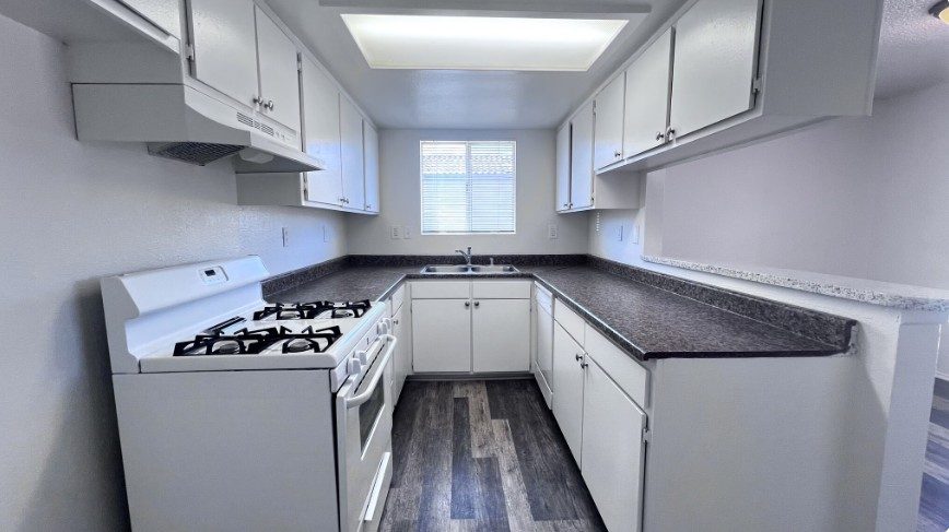 A renovated kitchen with white cabinets, a gas range, a double sink under the window, dark speckled countertops and grey wood-look plank flooring.
