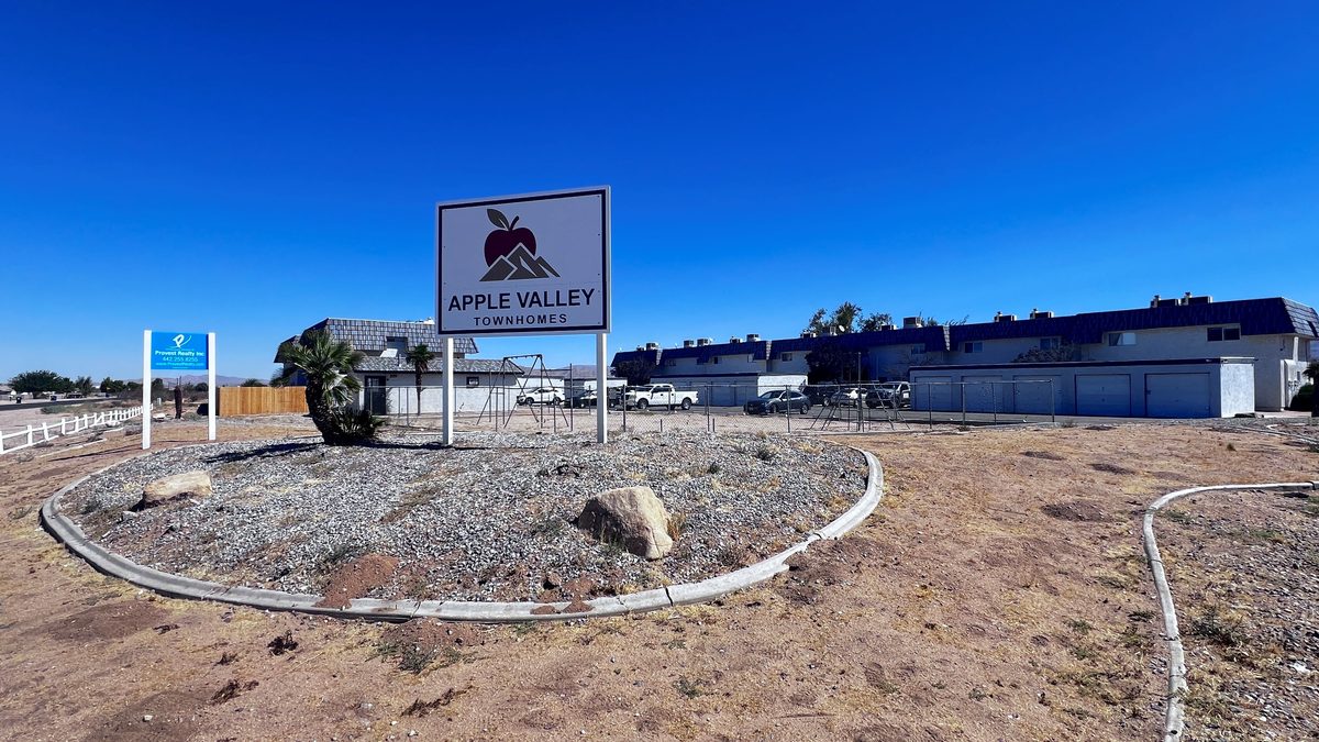 The Apple Valley Townhomes sign at the entrance on Sioux Road, the apple and mountain logo on a white panel against a deep blue sky, with the townhomes and garages behind it.