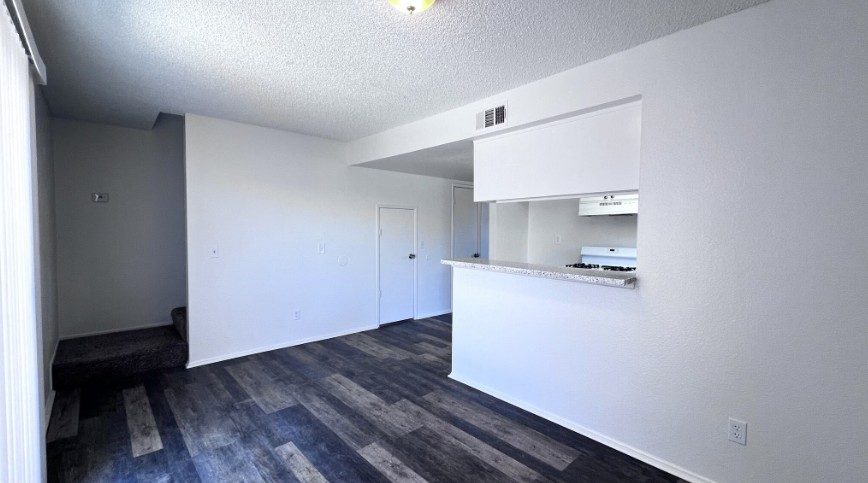 The living room of a renovated three bedroom townhome, grey plank flooring, white walls, and the pass-through counter to the kitchen, with the stairs to the bedrooms on the left.