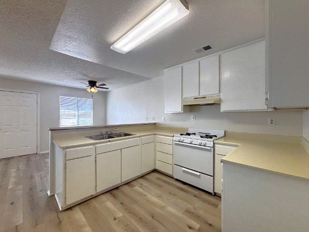 A renovated kitchen with white cabinets, a white gas range, a double sink under the window and light wood-look flooring, with a ceiling fan in the room beyond.