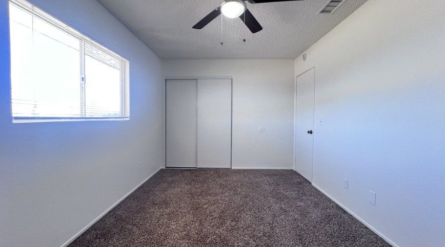 A bedroom with tan carpet, white walls, a ceiling fan, a wide window with blinds, and a sliding-door closet.