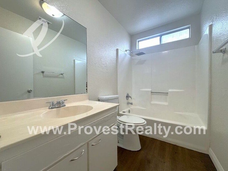 A bathroom with a tub and shower, a vanity with a white top and a wide mirror, and a window over the tub.