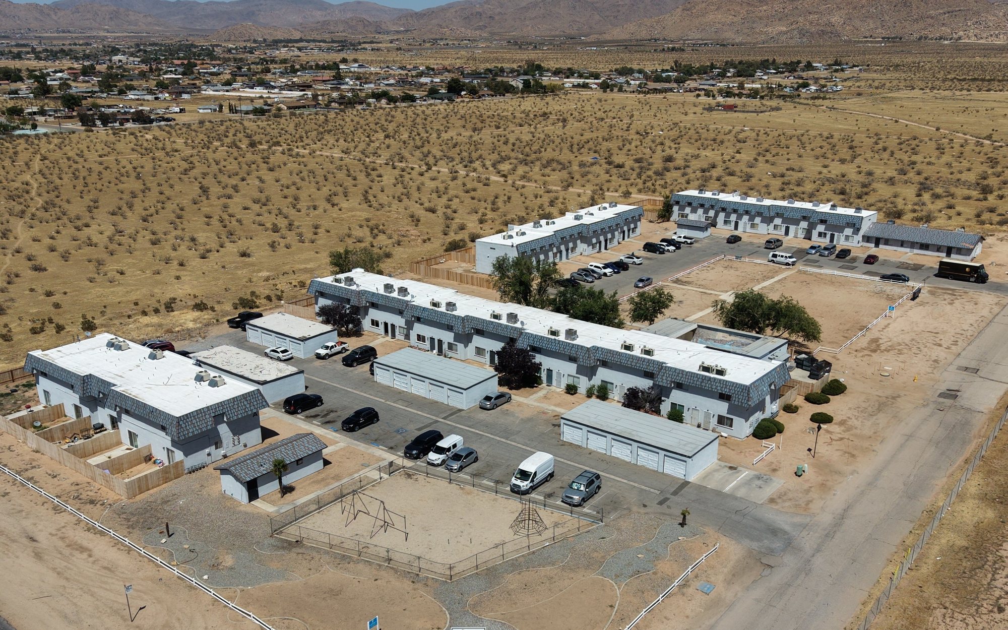 Aerial view of Apple Valley Townhomes from above Sioux Road: ten low two-story buildings with grey tile roofs around the parking lot, the garages and playground at the front, fenced backyards behind each row, the town spread out beyond and mountains along the horizon under a clear sky.